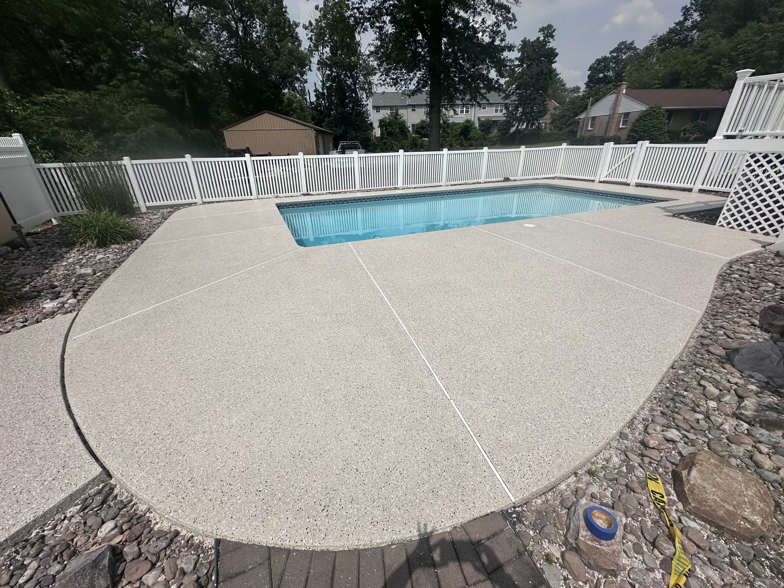 Cream flake pool deck with clean white saw cut control joints, an in ground pool and white vinyl fencing behind it