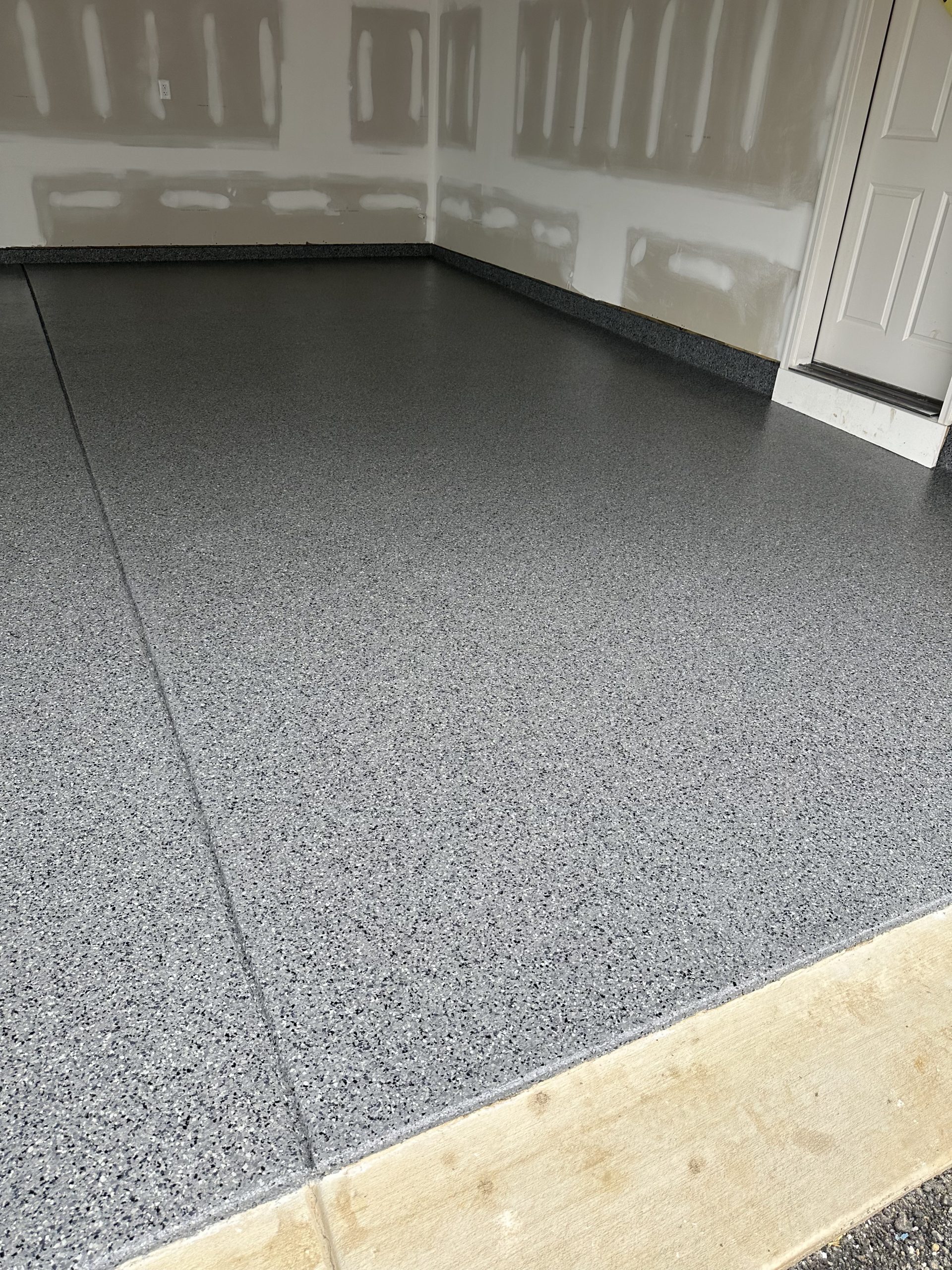 Gray flake floor with dark speckle running to unpainted drywall walls, a white door at the right and a raw timber threshold board across the front edge